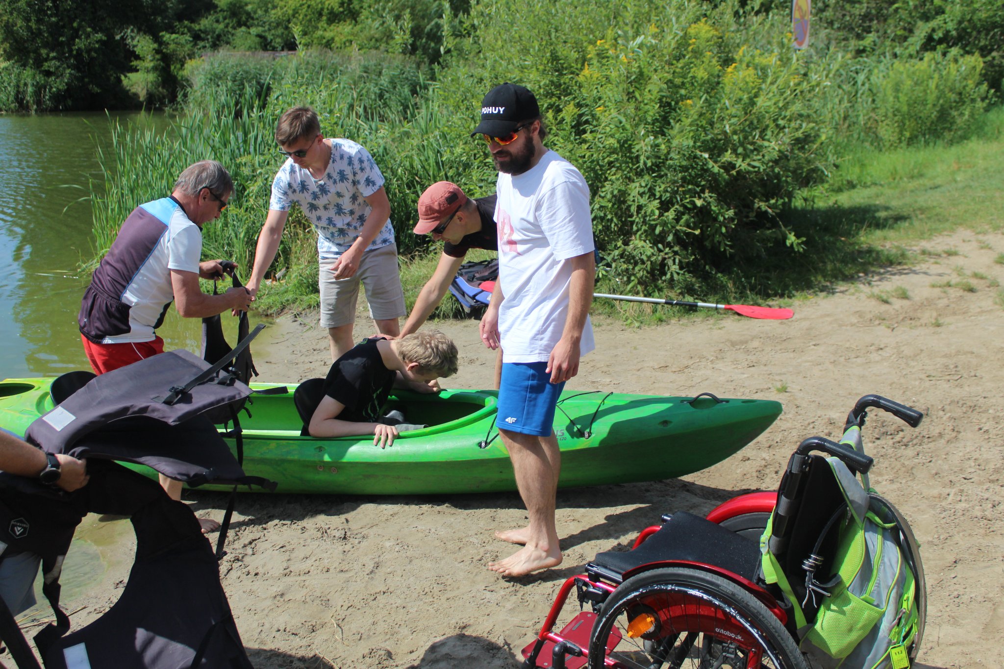 Grupa osób przebywająca na plaży, pomagająca wyjść niepełnosprawnemu chłopakowi z kajaku.