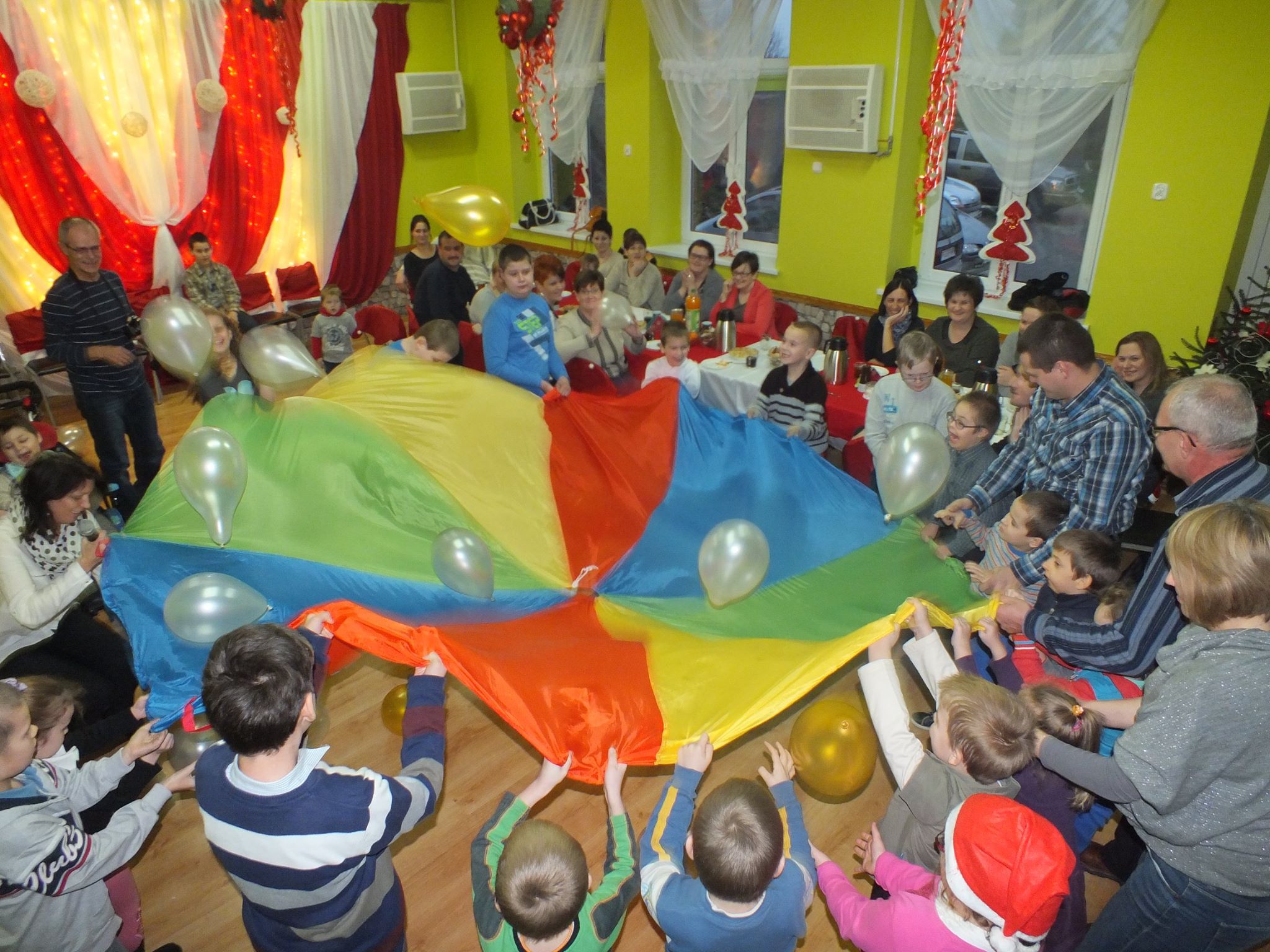 Grupa dzieci stojąca w okręgu trzyma kolorowy materiał i podbija nim balony. W tle siedzi grupa dorosłych przy stole i obserwuje dzieci.